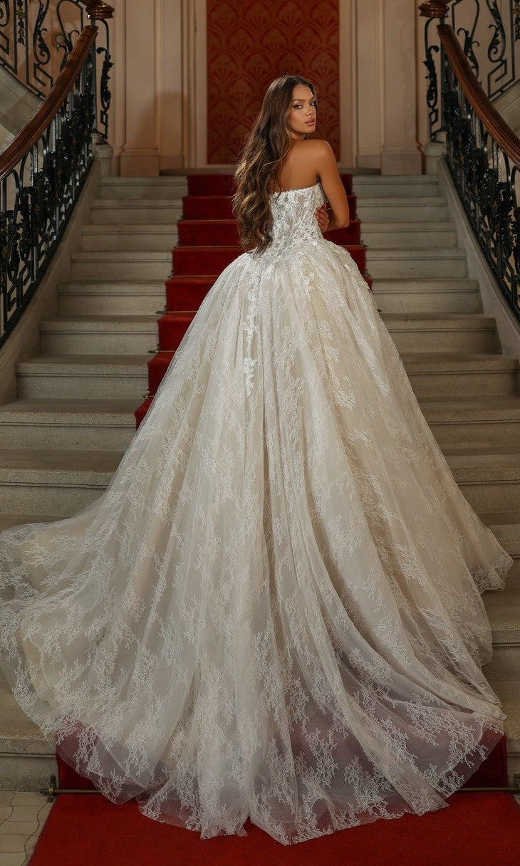 A woman in the Long White Dress: Alyce 7163 stands on a grand red-carpeted staircase, facing away and showcasing the strapless lace corset bodice.